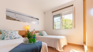 Une chambre d’hôtel avec deux lits, une fenêtre donnant sur un paysage verdoyant, une œuvre d’art accrochée au mur et une table de chevet avec une lampe et des objets décoratifs.