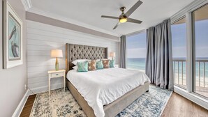 A bedroom with a large bed, a ceiling fan, a nightstand with a lamp, and a view of the ocean.