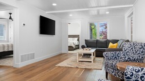 Ein geräumiges Wohnzimmer mit Holzdielen, einem wandmontierten Flachbildfernseher, einem grauen Sofa mit gelbem Kissen und einem gemusterten Sessel.