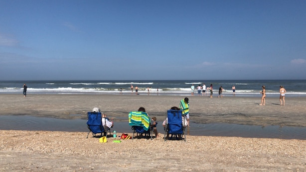 En strand med folk som promenerar och sitter på stolar, några av dem har strandparasoller.