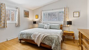 A bedroom with a large bed, two bedside tables, a wooden dresser, and two windows with curtains. - Bright "Mid-Century Modern" Ranch (Rochester)