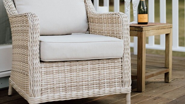 A wicker chair with a cushion on a wooden deck, a bottle of champagne, and a white railing.