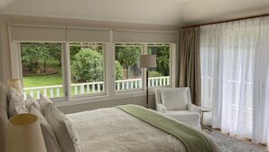 A bedroom with a large bed, a chair, a lamp, and a view of a garden through large windows.