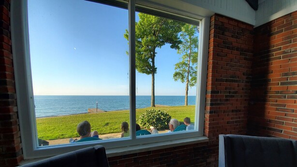 Ein Zimmer mit großem Fenster mit Blick auf eine Wiese und das Meer. Im Vordergrund stehen zwei Stühle.