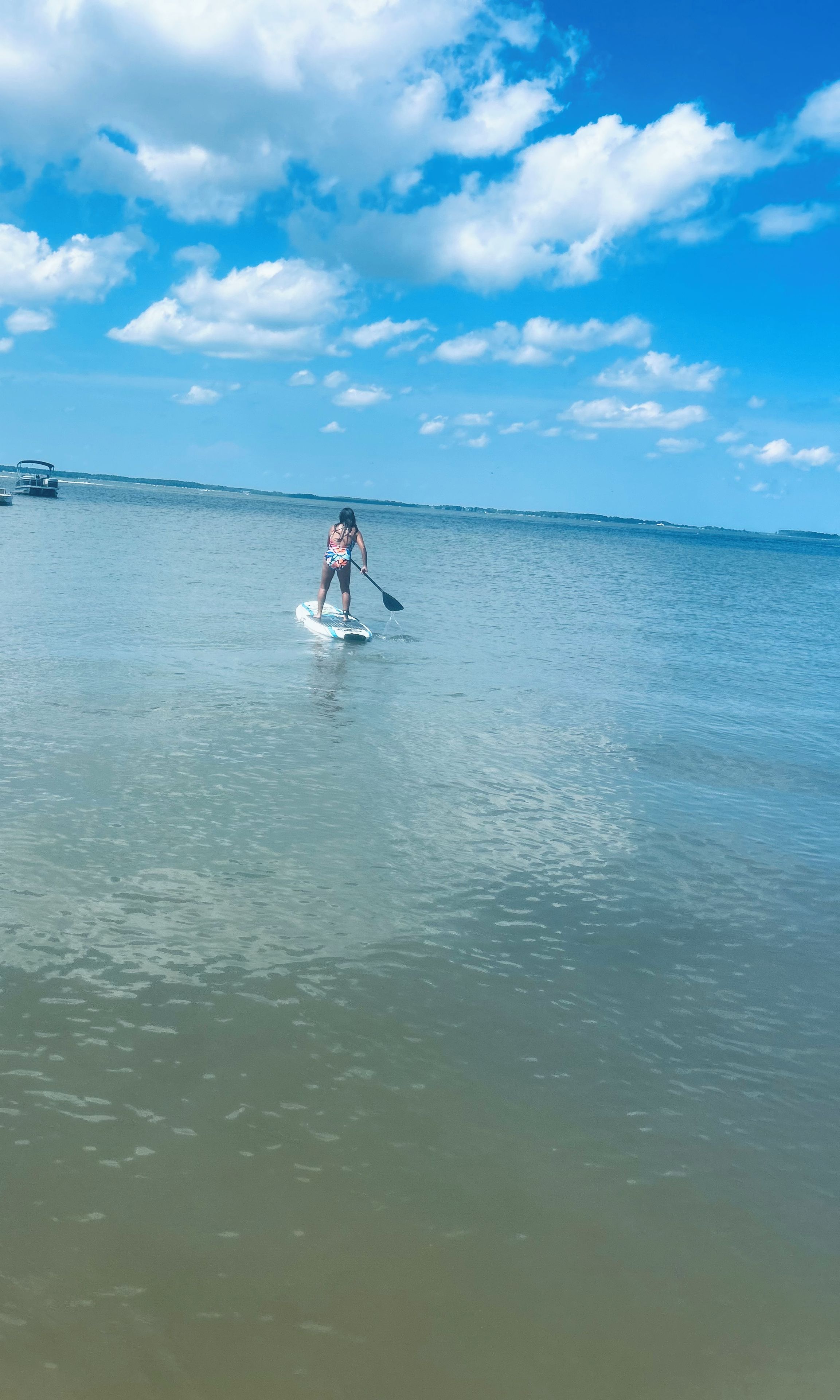 A person paddleboarding on calm water with a clear sky and distant land.