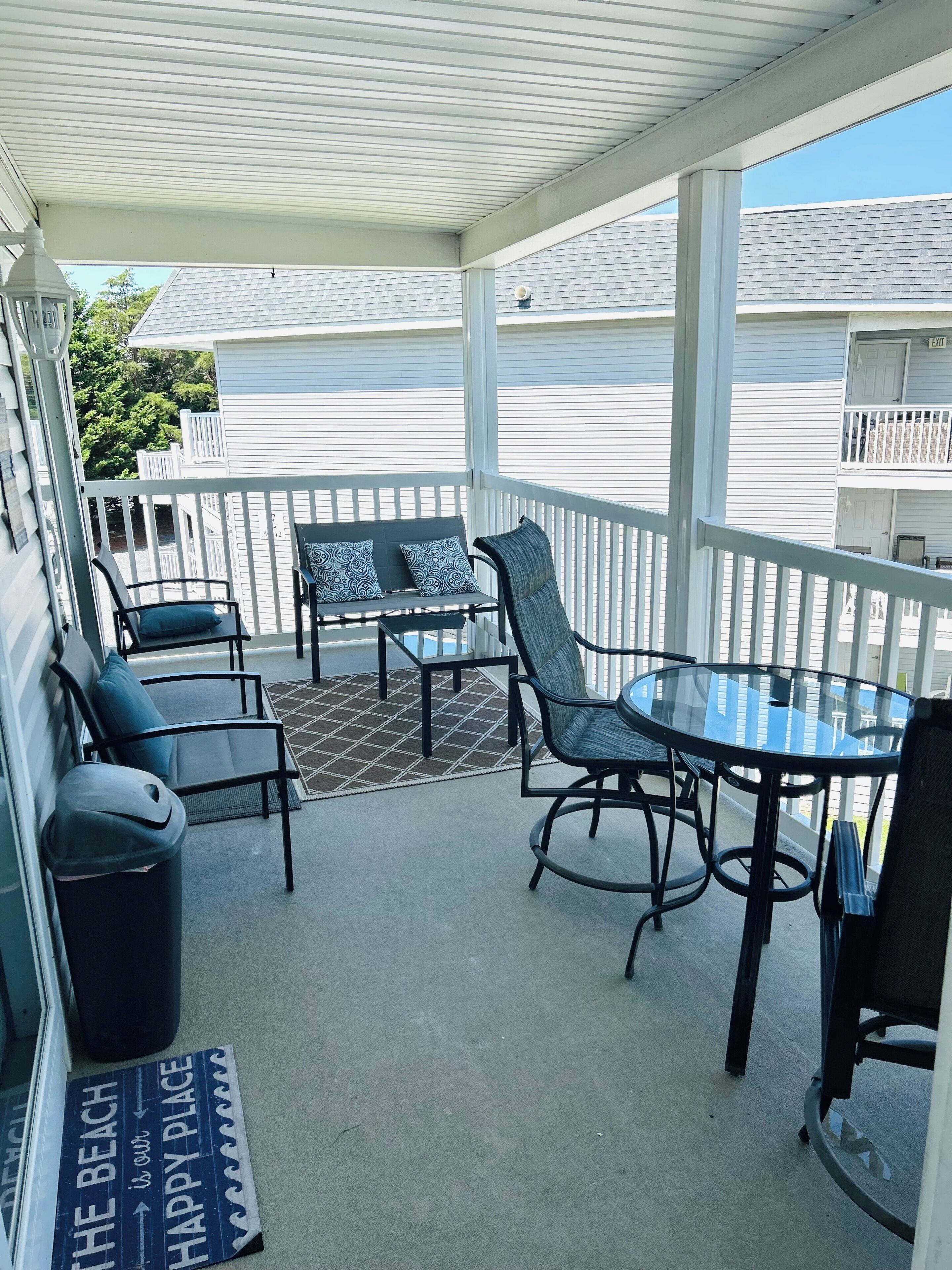 A covered outdoor seating area with a glass table and chairs, a trash can, and a mat with text.