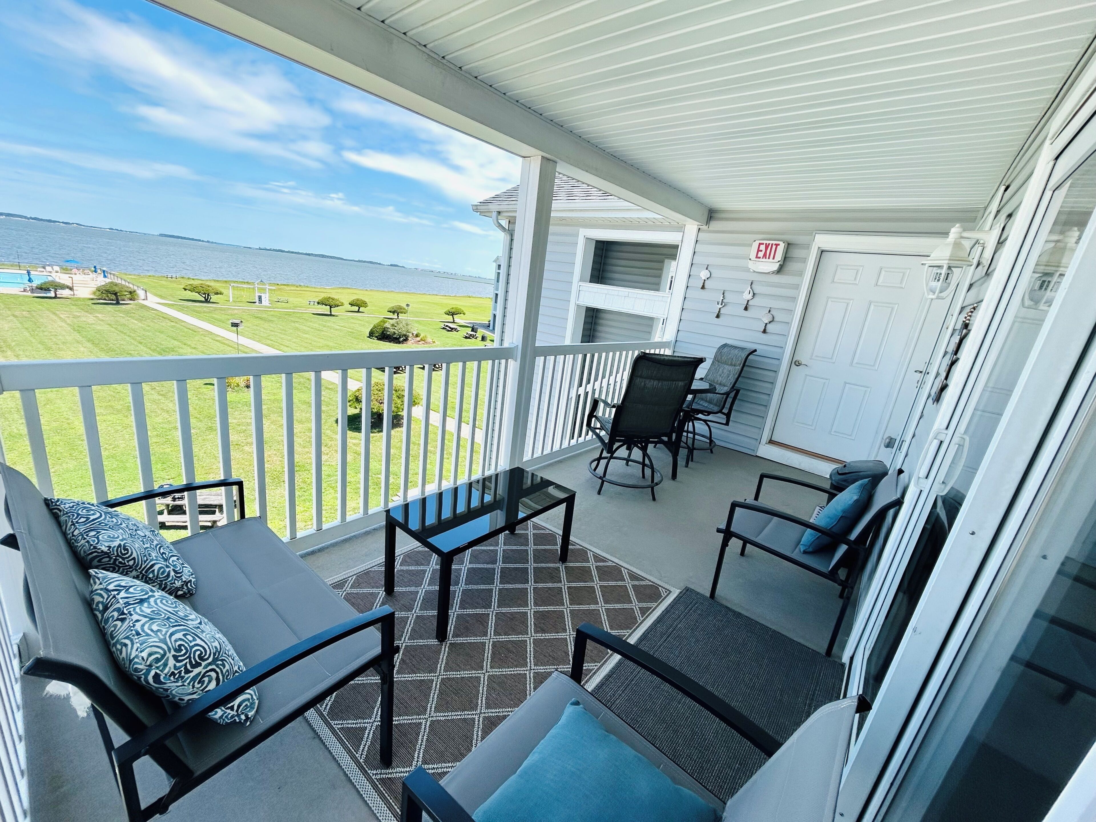 A covered balcony with a view of the water, a table with chairs, and a door with an exit sign.