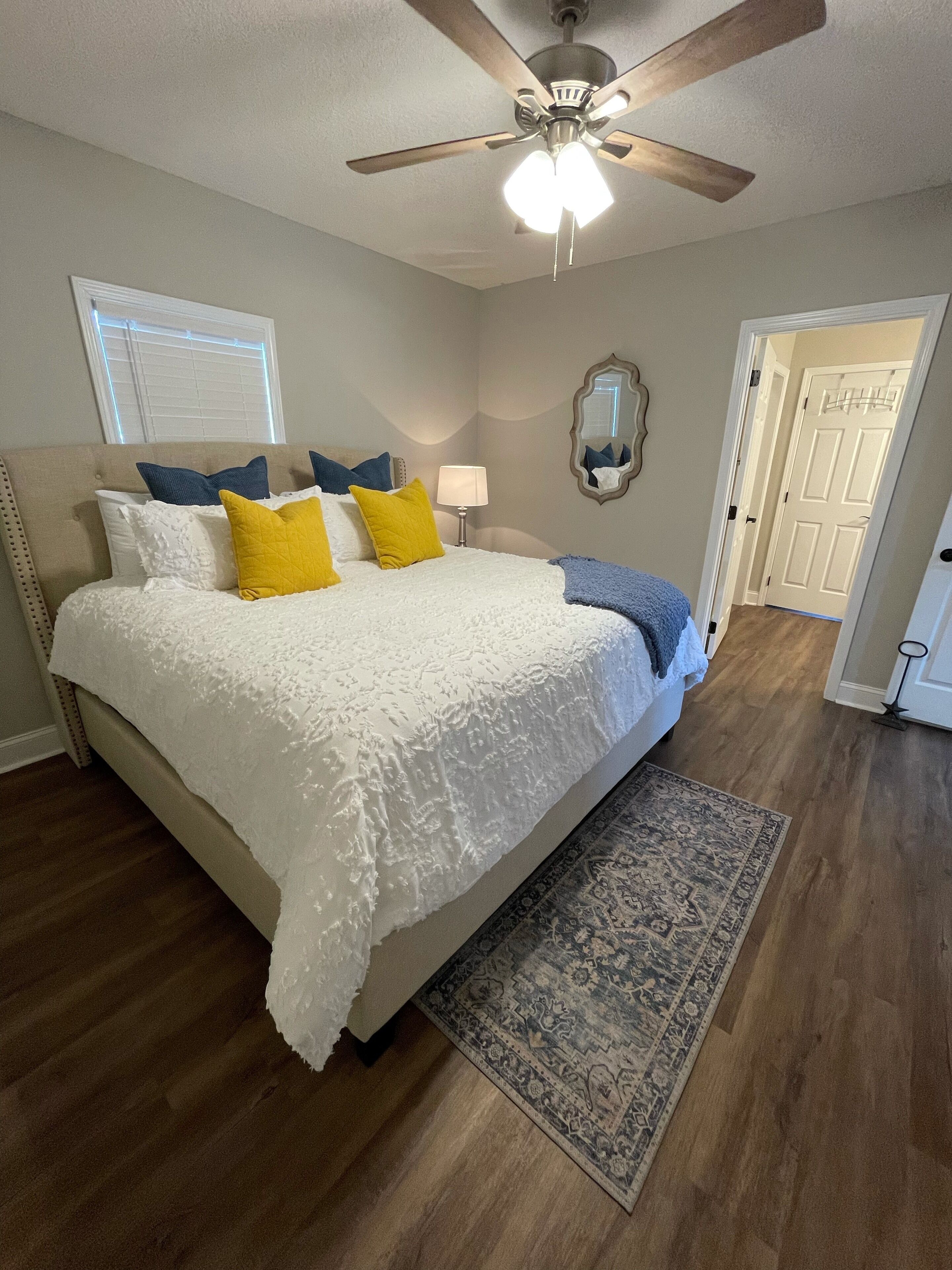 A bedroom with a bed, a ceiling fan, a nightstand, a lamp, a mirror, and a door.