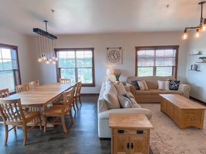 Amplia sala de estar con una mesa de comedor de madera, un sofá y una mesa de centro. Hay ventanas grandes con persianas, un candelabro en el techo y un reloj de pared.