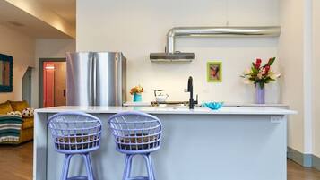 A modern kitchen with a central island, two bar stools, a stainless steel refrigerator, a sink, and a decorative vase.
