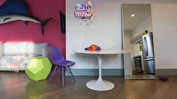 Un salón moderno con una mesa redonda blanca, sillas moradas, una manta tejida de colores, un otomano hexagonal verde, un pez colgado en la pared y un letrero de neón que dice "TIKIBAR".
