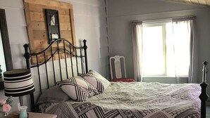 Ett sovrum med ett träpanelat tak, en säng med mönstrat sänglinne, ett nattduksbord med en lampa och ett fönster med gardiner.