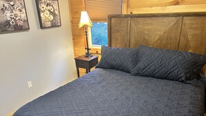 A bedroom with a large bed, a nightstand with a lamp, and wooden paneling on the walls. - Cozy cottage condo with easy access to TIEC, Hendersonville, Asheville & Boone (Rutherfordton)