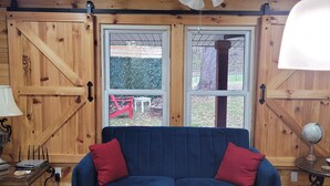 A cozy room with a wooden ceiling, a ceiling fan, a blue sofa with red cushions, a wooden coffee table, and a view of a fenced yard through large windows. - Cozy cottage condo with easy access to TIEC, Hendersonville, Asheville & Boone (Rutherfordton)