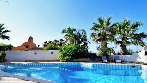 A swimming pool with blue water, surrounded by palm trees and a white building with a unique architectural design. - Le Palme by Interhome (Valderice)
