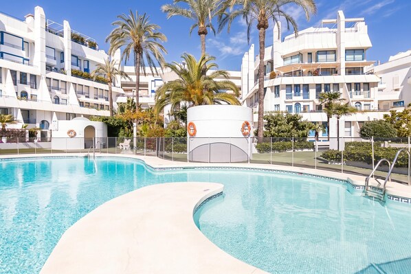 A swimming pool with a curved edge, surrounded by palm trees and modern buildings with balconies.
