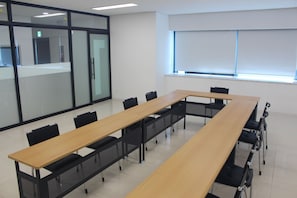 Conference room with a long table and black chairs, featuring a whiteboard and large window with blinds.