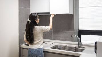 Una mujer en una cocina con un lavamanos de acero inoxidable, una ventana con persianas y una encimera.