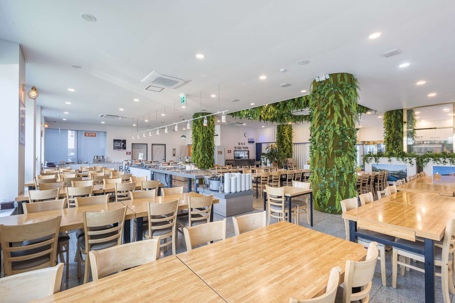 Uma cafeteria ampla com mesas e cadeiras de madeira, uma área central de serviço e jardins verticais verdes.