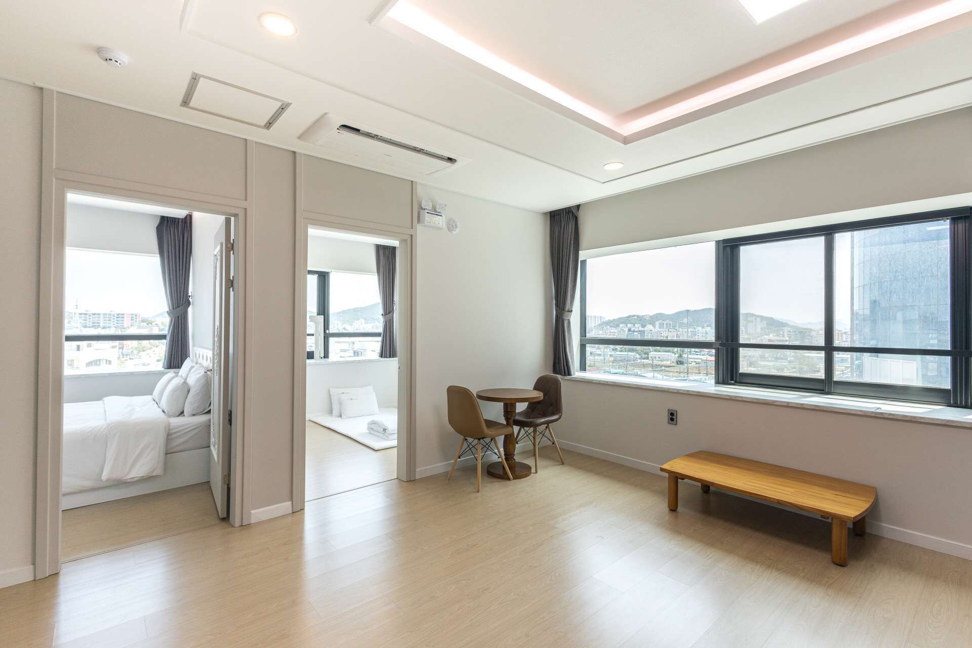 Habitación de hotel con cama, una pequeña mesa con sillas, un banco y un amplio ventanal con vistas a la ciudad.