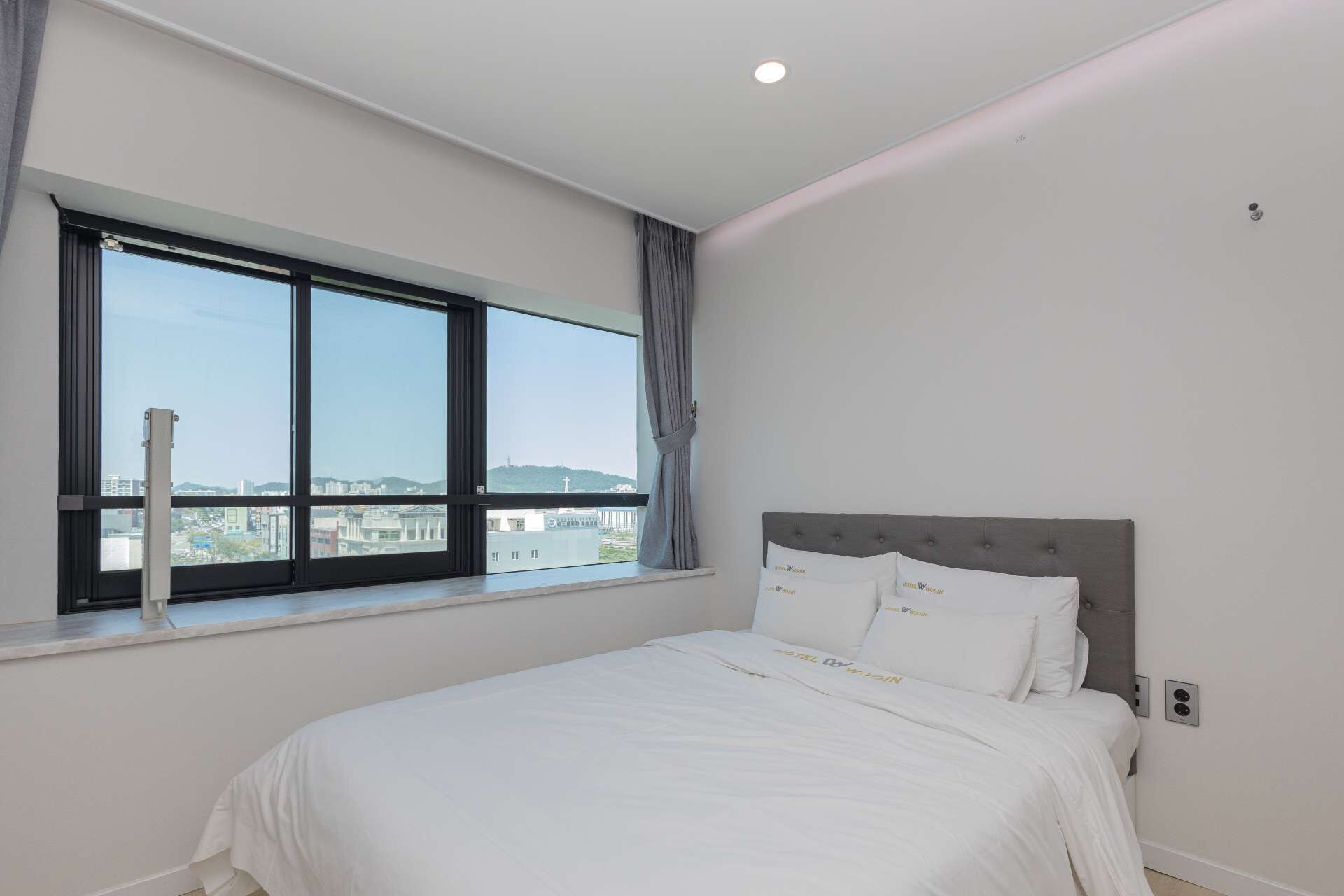 Un dormitorio con un ventanal grande, una cama con ropa de cama blanca y vistas a un paisaje urbano.