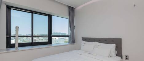Un dormitorio con un ventanal grande, una cama con ropa de cama blanca y vistas a un paisaje urbano.