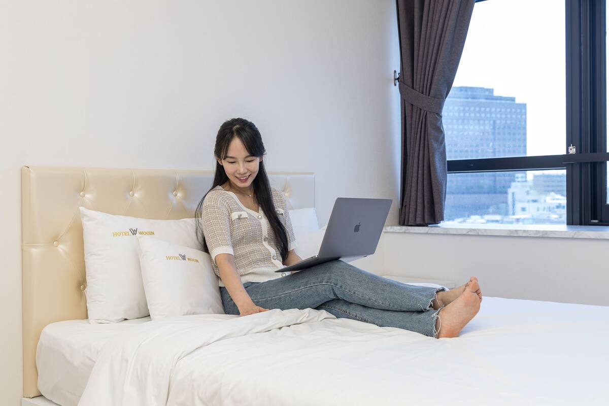 A person sitting on a bed with a laptop, a window with curtains, and pillows with hotel branding.
