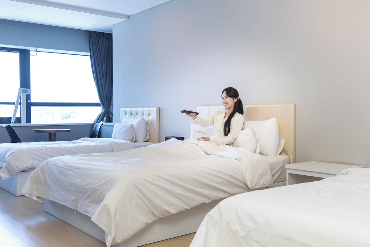 A hotel room with two beds, a bedside table, a chair, a window with curtains, and a view of the city.