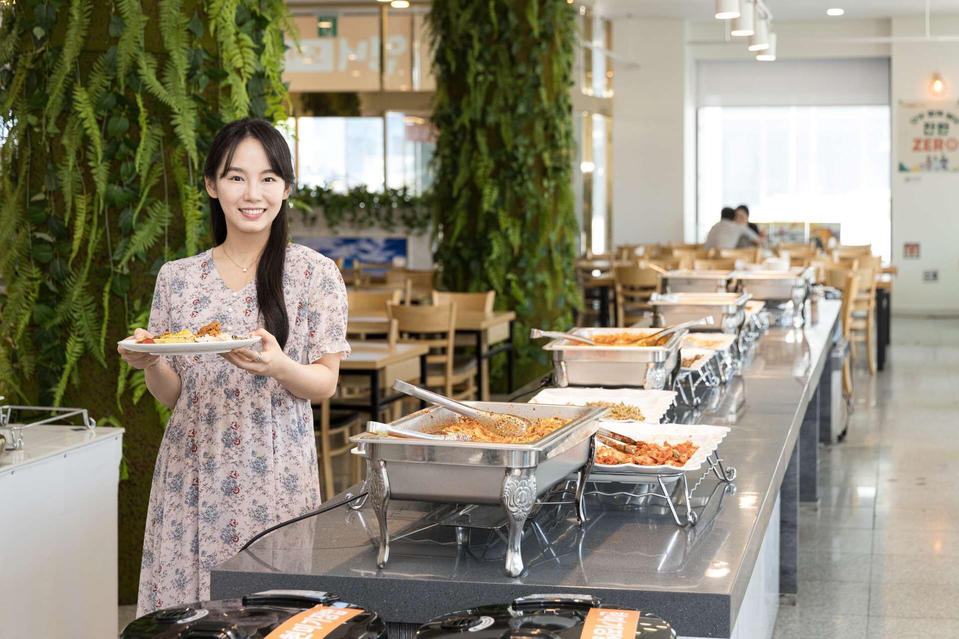 A buffet setup with multiple serving stations, a person holding a plate, and a modern interior with plants and seating areas.