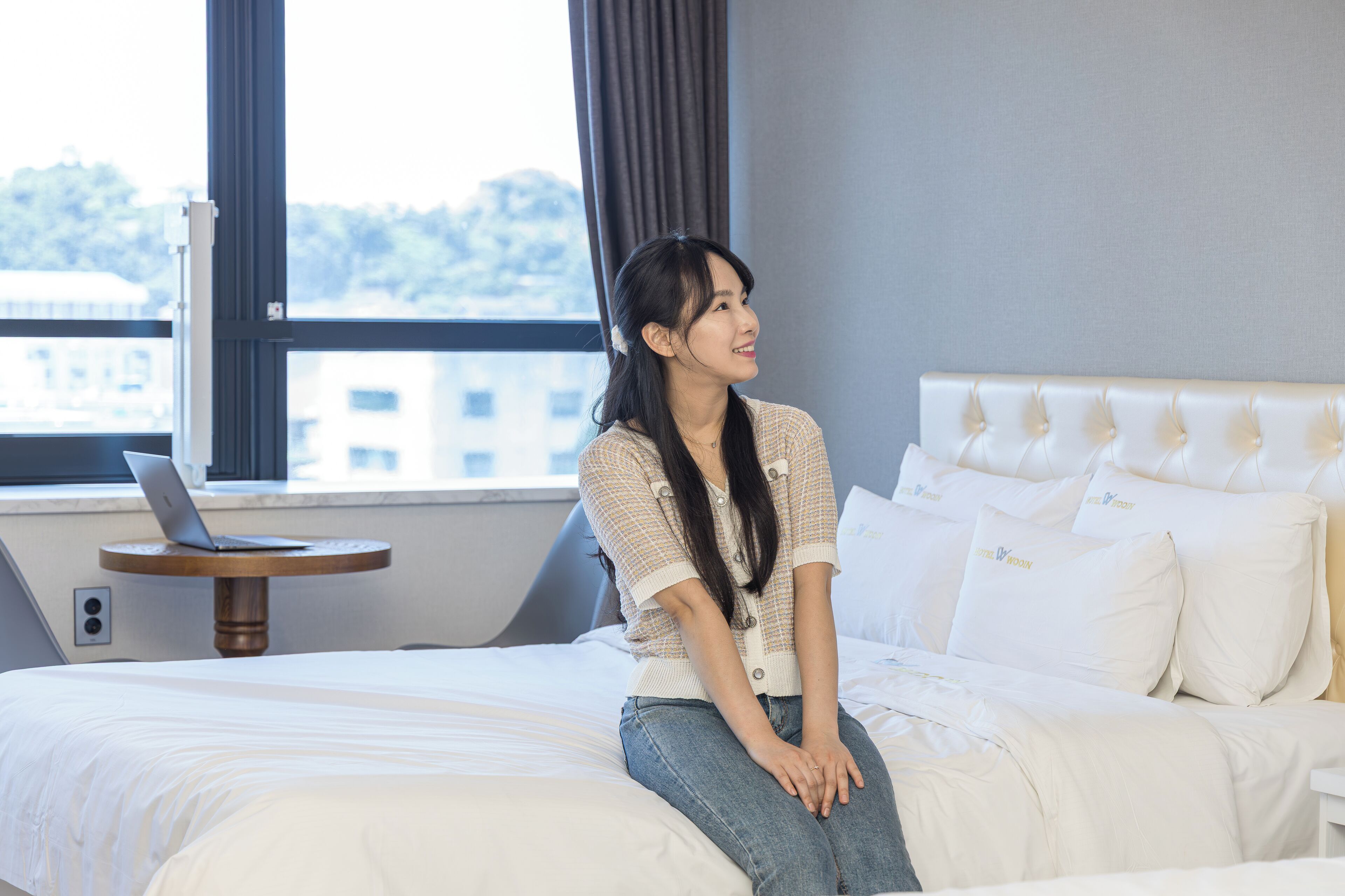 Una habitación de hotel con una cama, una computadora portátil sobre una mesita y una ventana con vistas a edificios.