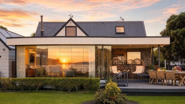 Ein modernes Haus mit großen Glasfenstern, einer Terrasse mit Korbmöbeln und einem gepflegten Rasen.