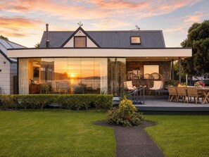 A modern house with large glass windows, a patio with wicker furniture, and a well-maintained lawn. - Blackpool Bayside - Spa - Easy Walk to Oneroa (Oneroa)