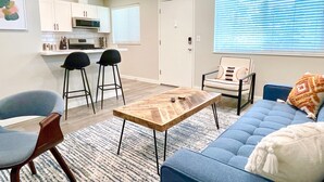 Een moderne woonkamer met een blauwe bank, een salontafel en twee barkrukken. Er is een keuken met witte kastjes en een magnetron.