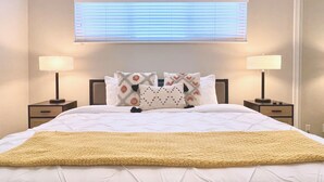 Een net opgemaakt bed met een witte deken en een gele plaid, met aan beide zijden nachtkastjes en lampen, een raam met zonwering, en een hoofdbord met decoratieve kussens.