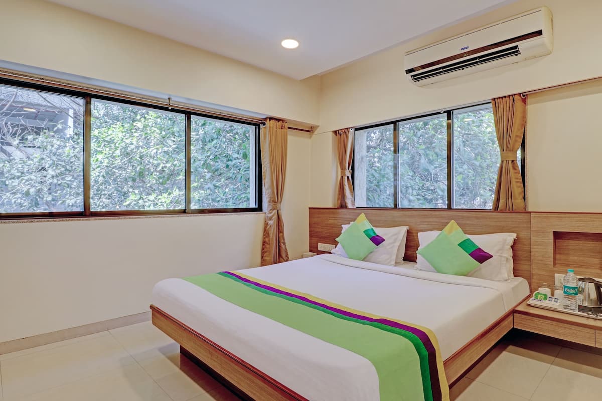 Habitación de hotel con una cama grande, dos ventanas con cortinas, un equipo de aire acondicionado y una mesita de noche con una bandeja que contiene una tetera, una botella de agua y otros objetos.