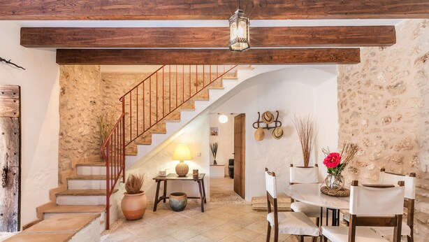 A rustic interior with a staircase, wooden beams, a dining area with chairs and a table, and a stone wall.