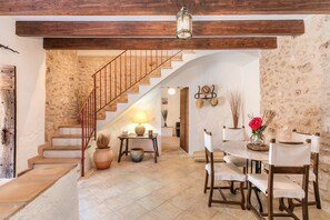 Un intérieur rustique avec un escalier, des poutres en bois, un coin repas avec des chaises et une table, et un mur en pierre.