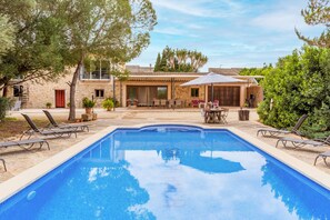 Une piscine avec des chaises longues, un patio couvert et un bâtiment en pierre surmonté d’un toit en tuiles.