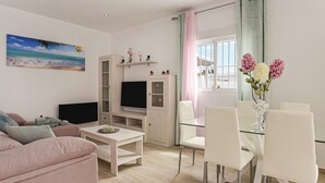 Ein modernes Wohnzimmer mit Flachbildfernseher, einem Glastisch, weißen Stühlen, einem rosafarbenen Sofa und einem Strandbild.