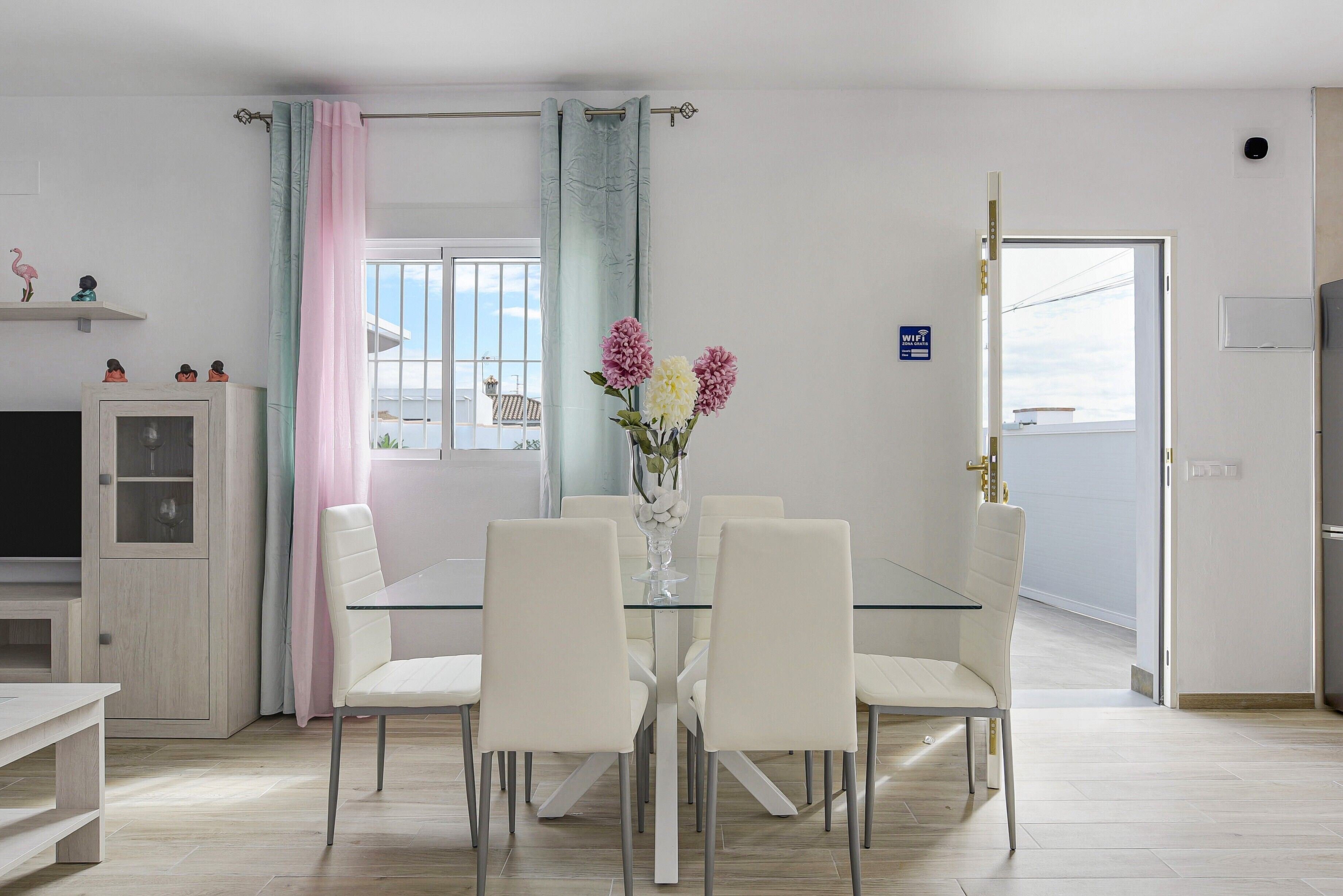 A dining area with a glass table and white chairs, a vase with flowers, and a view through the window.