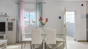 A dining area with a glass table and white chairs, a vase with flowers, and a view through the window.