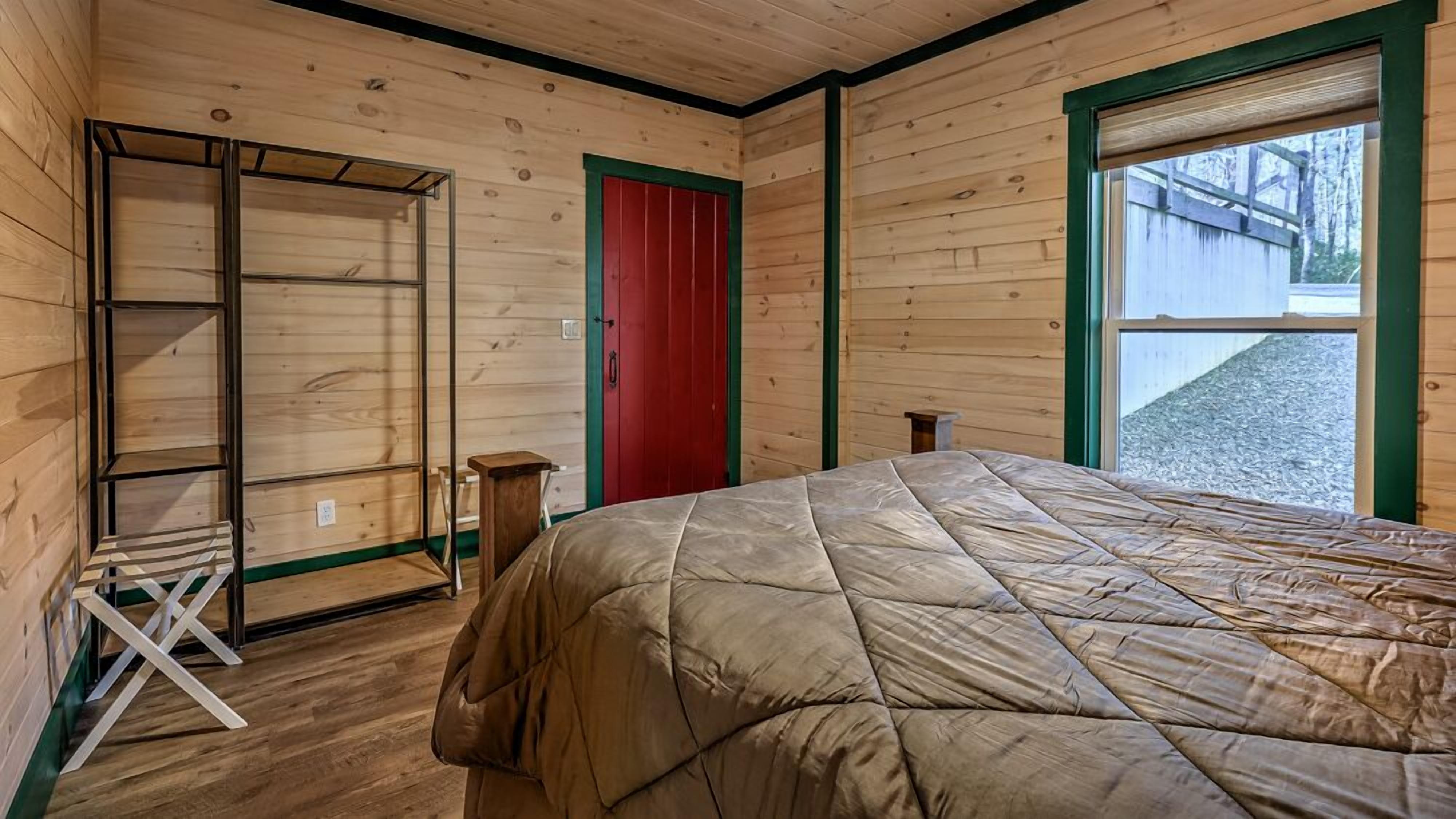 A wooden room with a bed, a red door, and a window.