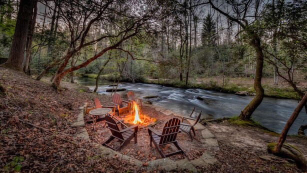 Ein Campingplatz mit Feuerstelle und Stühlen an einem Fluss in einem Wald.