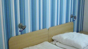 Een bed met witte beddengoed, een houten hoofdbord en een nachtkastje met een lade. De kamer heeft blauw-wit gestreept behang.