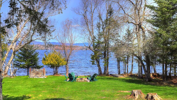 A lakeside property with a grassy lawn, two Adirondack chairs, and a wooden deck. - Niboban Cabin 5 right on Rangeley Lake with a screened porch, fire pit, hammock, WiFi and dock! (Rangeley)