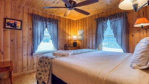 A wooden cabin bedroom with a bed, a ceiling fan, a nightstand, and two windows with checkered curtains. - Niboban Cabin 5 right on Rangeley Lake with a screened porch, fire pit, hammock, WiFi and dock! (Rangeley)