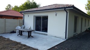 Un bâtiment d’un seul étage, doté d’un toit en tuiles, d’une grande porte-fenêtre coulissante et d’une terrasse en béton avec une table et des chaises.