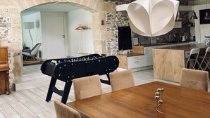 A dining area with a wooden table, cushioned chairs, a foosball table, and a stone wall.