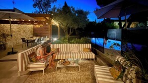An outdoor patio with a pool, a stone wall, and a wooden pergola with string lights.