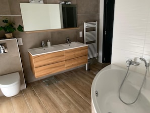 Een moderne badkamer met een houten wastafel, een wit toilet, een grote spiegel en een bad met douchekop.